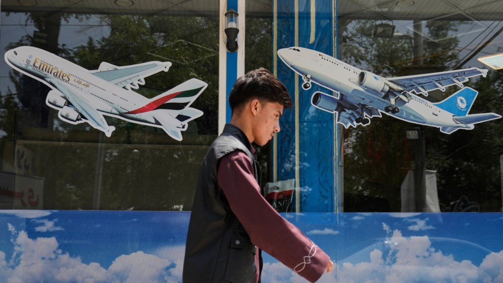 A young man in dark red top and black vest, shown from the waist up, walks by a shopfront decorated with images of airplanes and a blue sky with clouds.